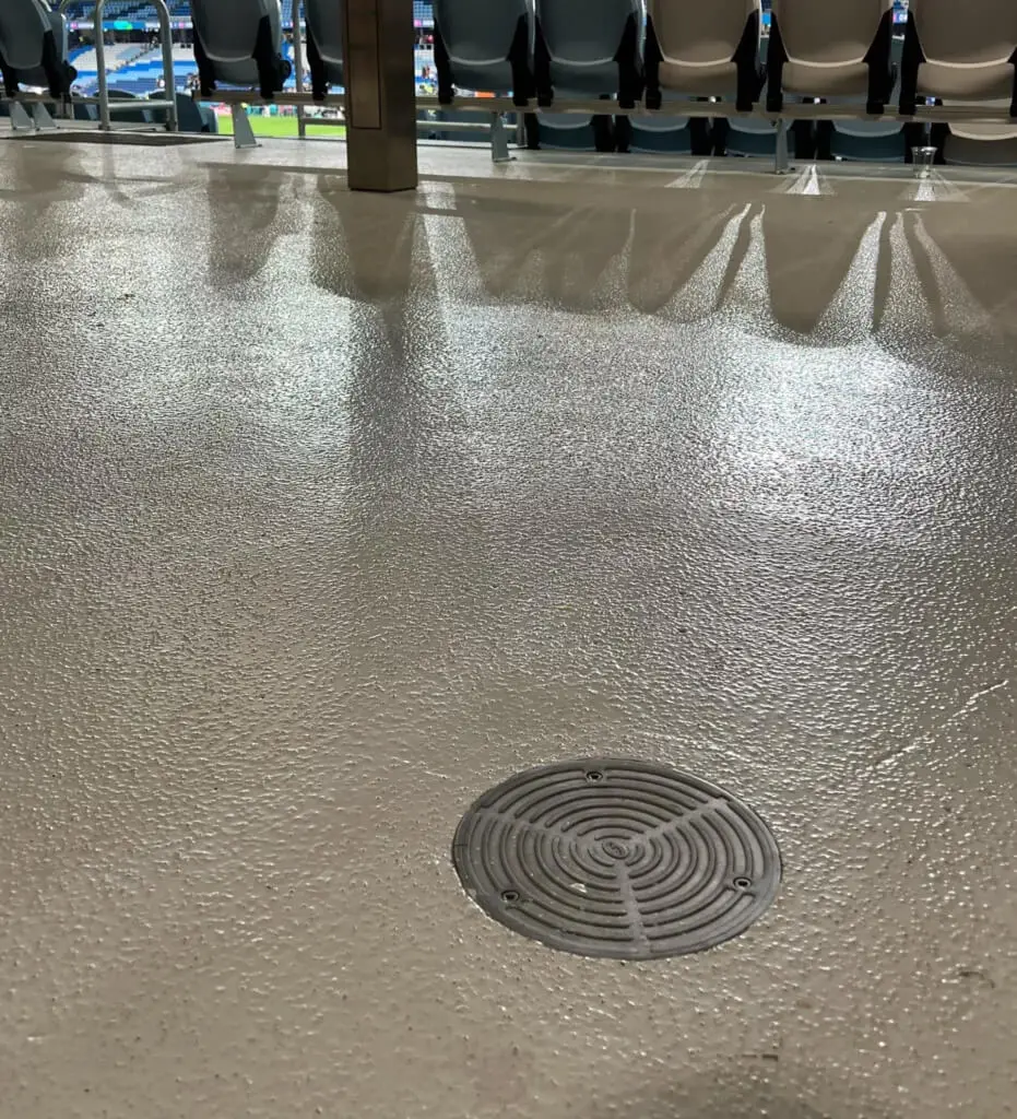 A close-up of a floor drain on a textured surface in front of a row of empty stadium seats, with a sports field visible in the background.