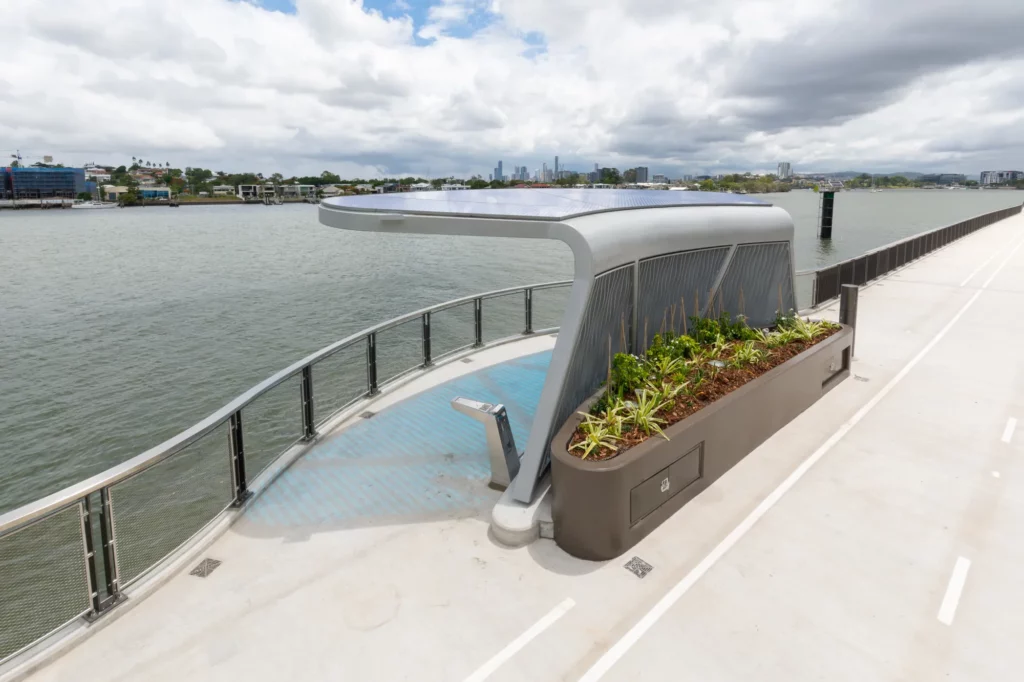 Riverside view featuring a modern pedestrian walkway with a sleek bench and a planter. The structure includes solar panels on a canopy, and the skyline is visible under a cloudy sky.